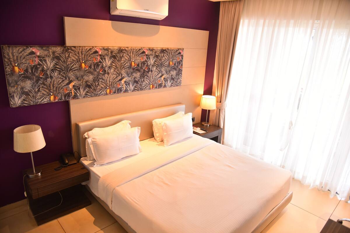 A hotel room with a large bed, two white pillows, a nightstand with a lamp, a phone, and a wall art piece above the bed.