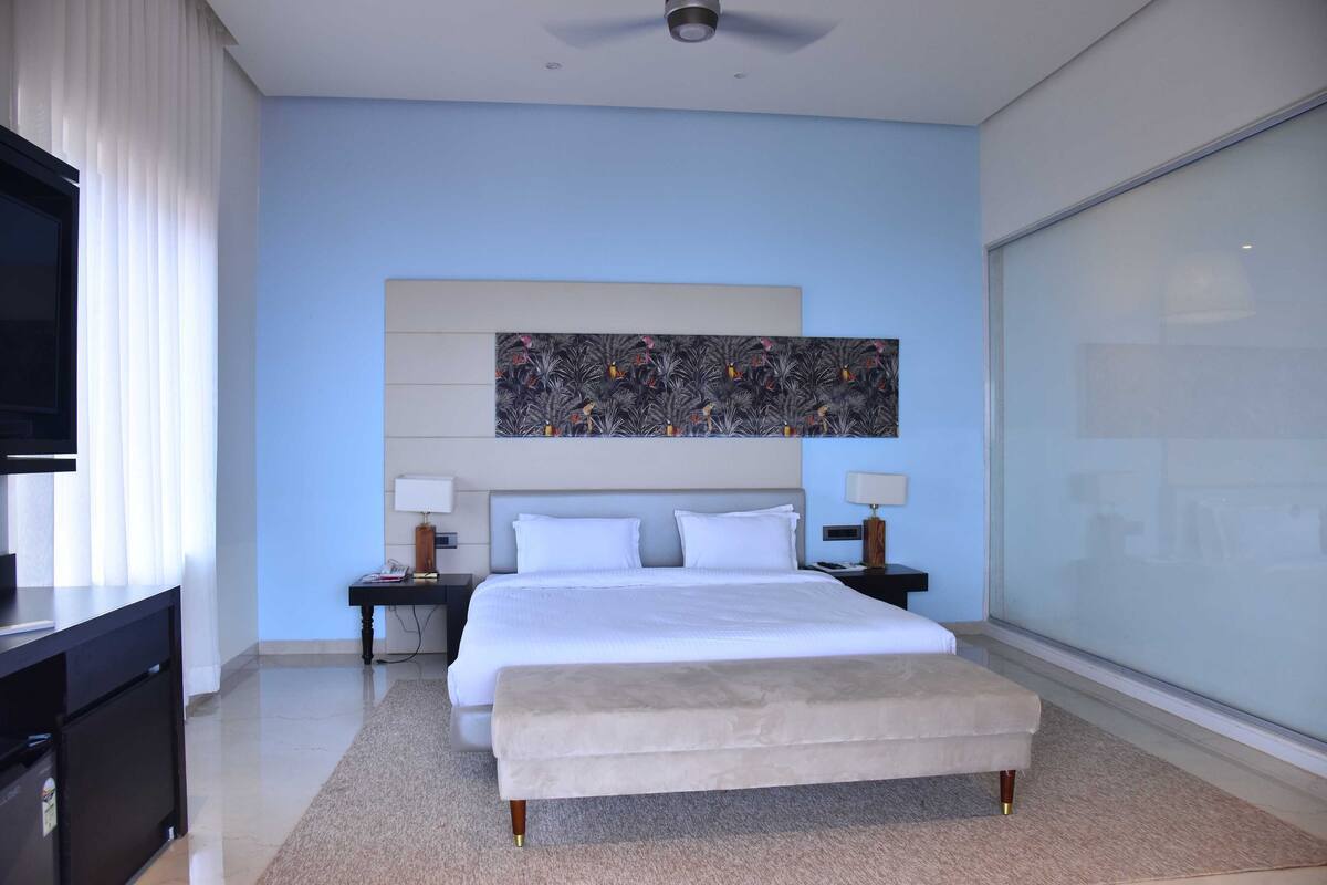 A modern hotel room with a large bed, bedside tables with lamps, a flat-screen TV, and a large window.