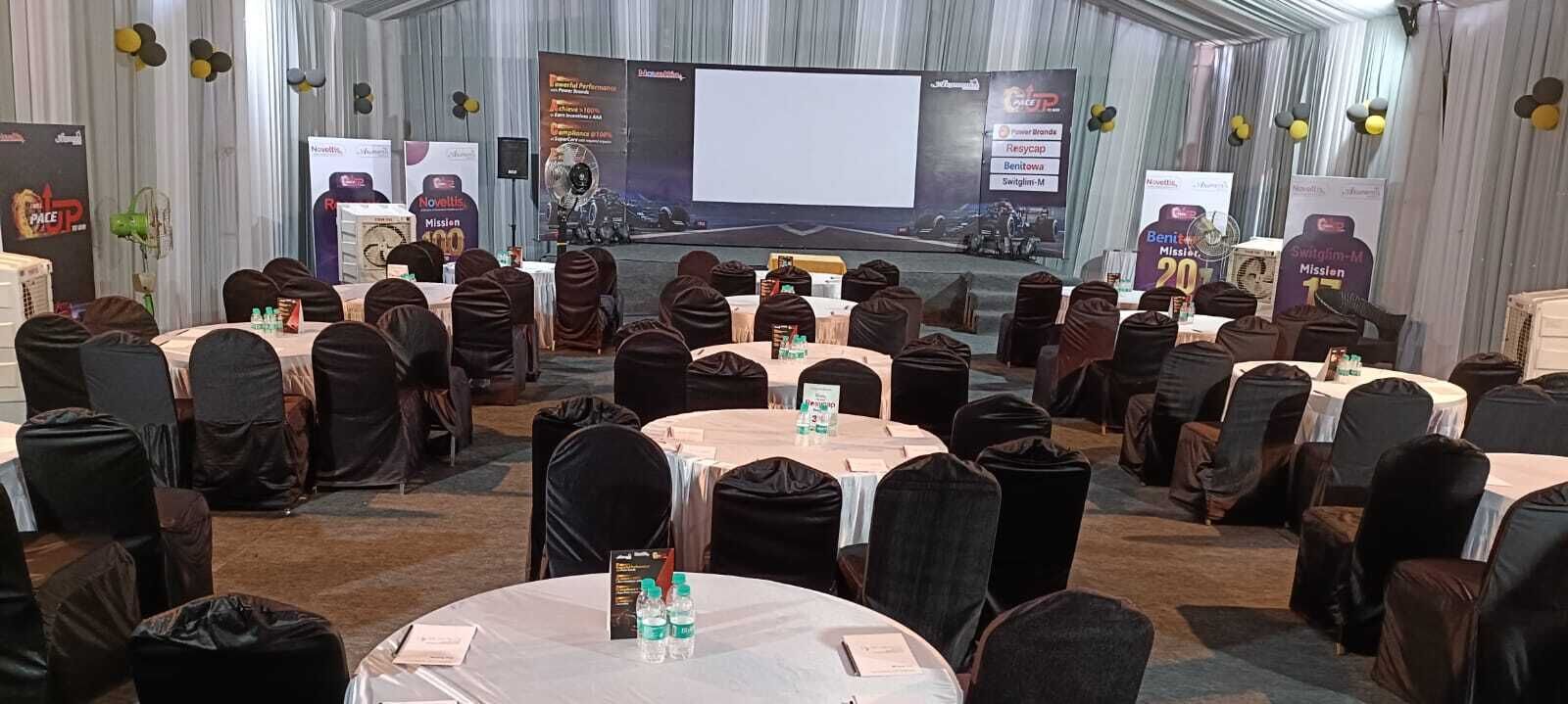 Une salle de conférence équipée de tables rondes, de chaises et d’une scène avec un écran de présentation.