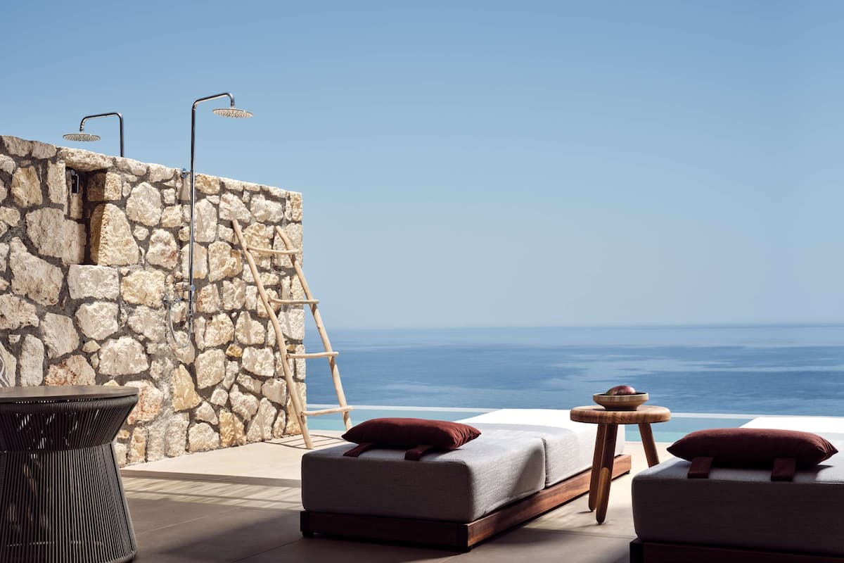 Una pared de piedra con una escalera, una mesa de madera, dos sillas con cojines y una cama con almohadas.
