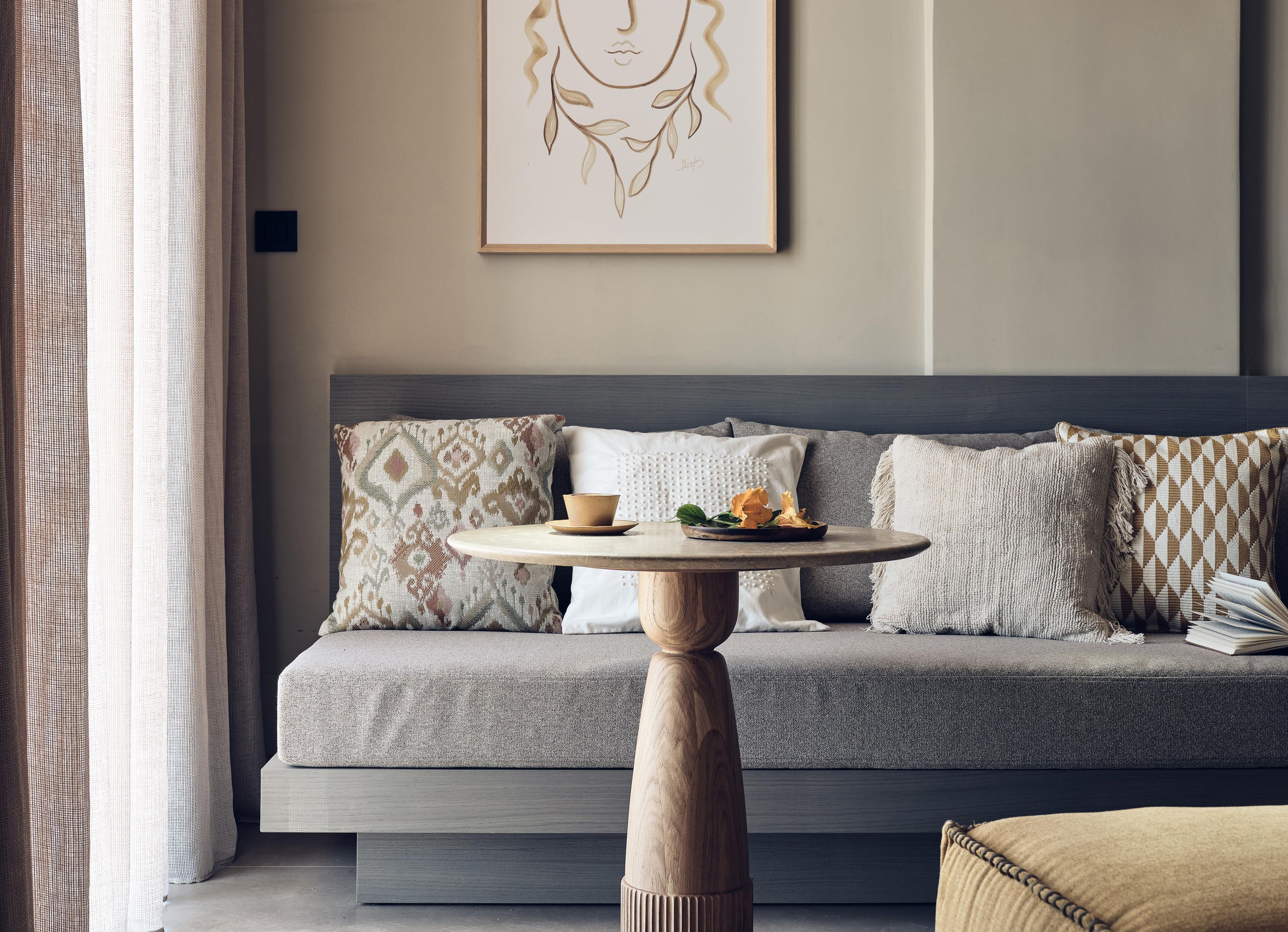A modern living room with a grey sofa, patterned cushions, a round wooden table with a bowl and flowers, and a framed artwork on the wall.