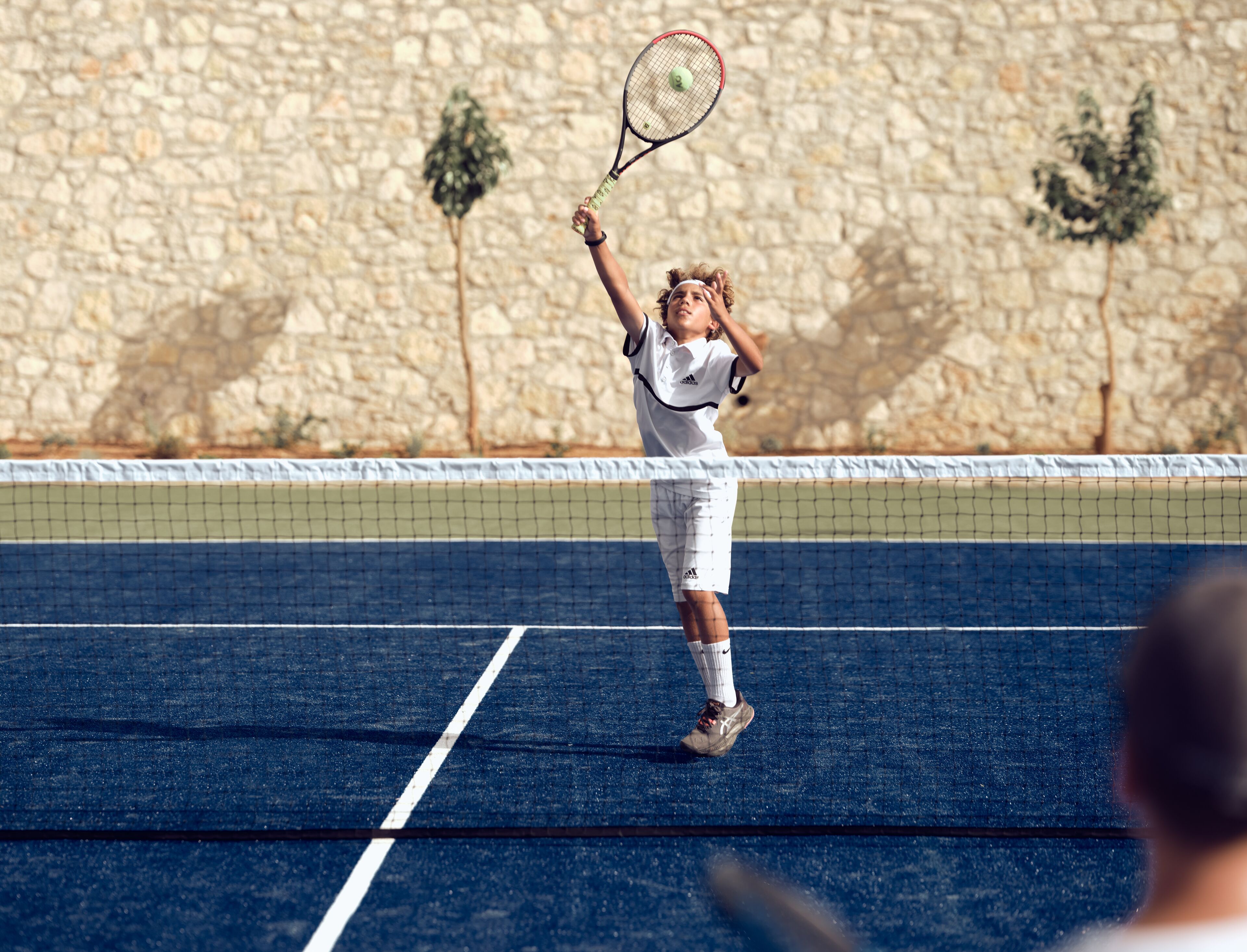 A tennis player in mid-action on a blue court with a stone wall in the background.