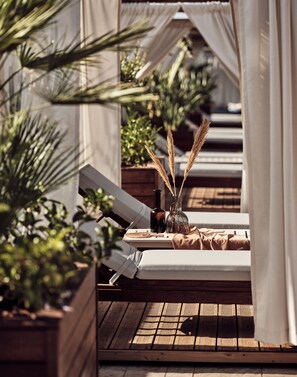 Un espacio lounge al aire libre con luz solar, suelo de madera, asientos acolchados y plantas en macetas.