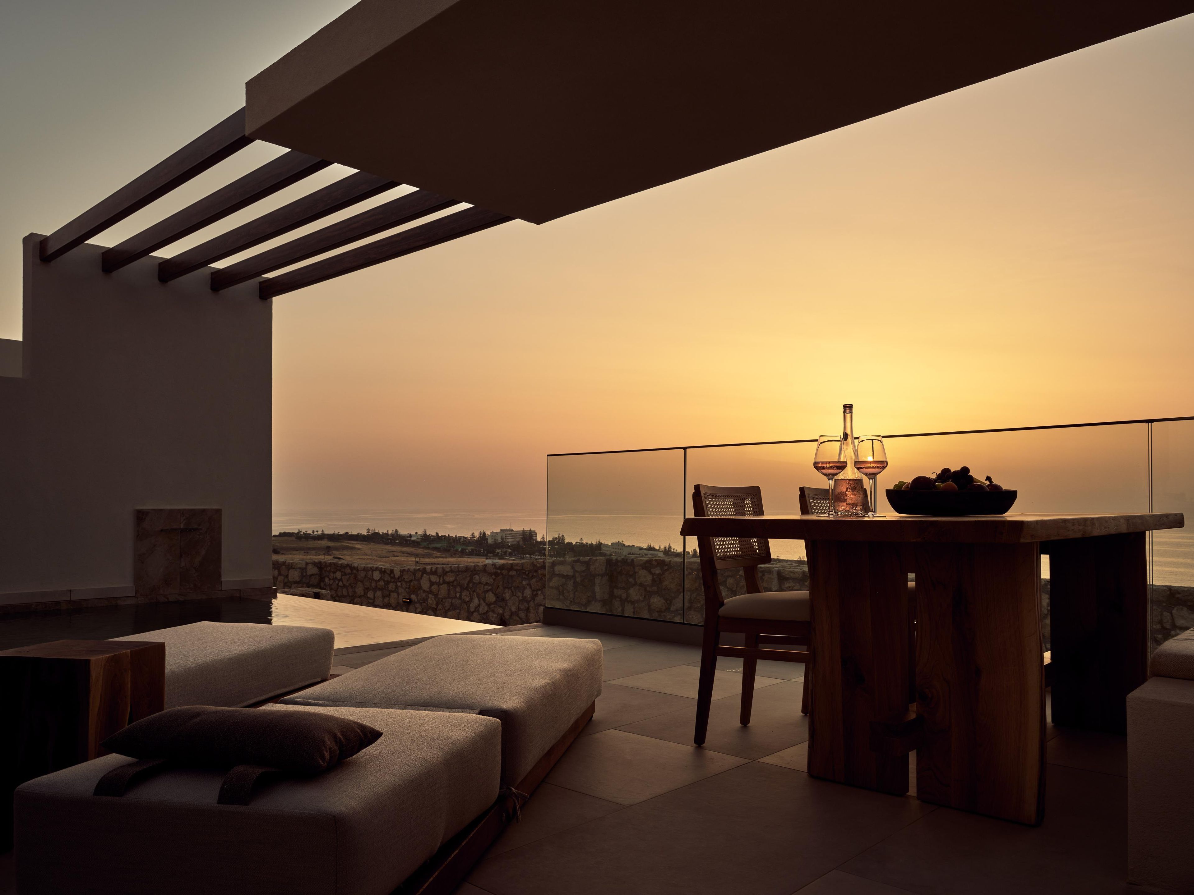 Un área de estar moderna al aire libre con una mesa, sillas y vistas al mar al atardecer.