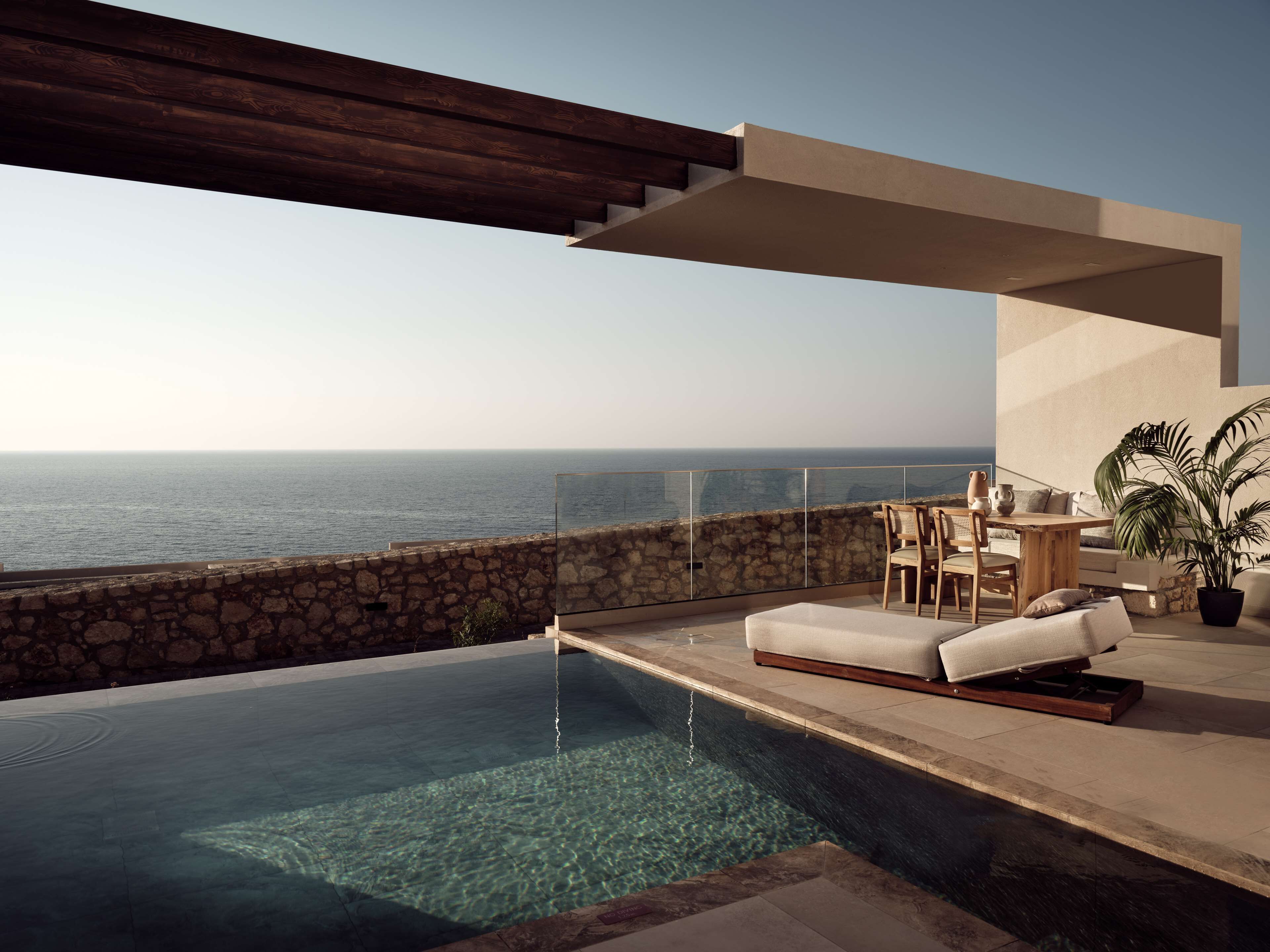 A modern outdoor pool area with a stone wall, wooden deck, and a view of the ocean.