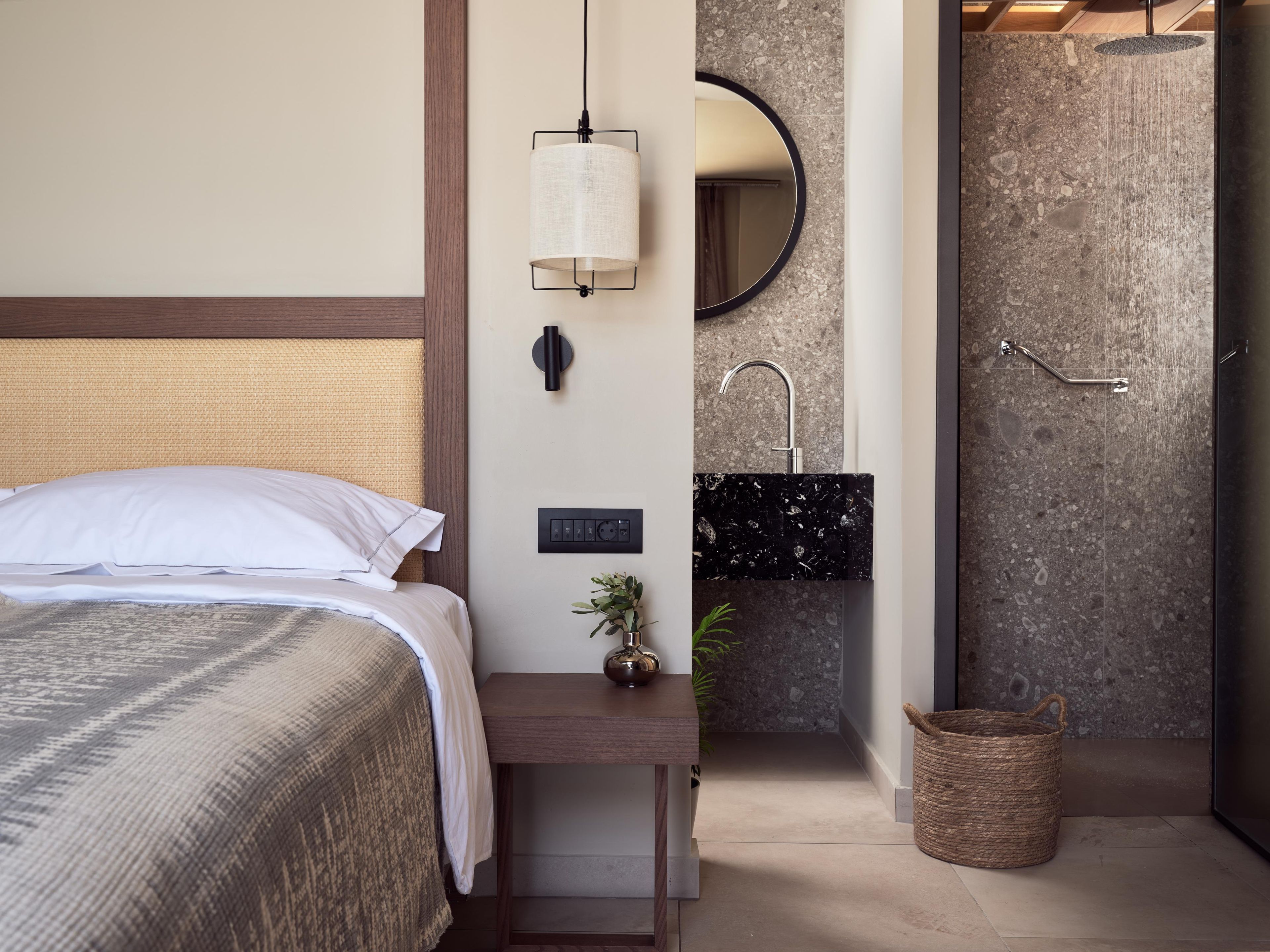 A hotel room with a bed, bedside table, lamp, wall-mounted mirror, sink, and shower.