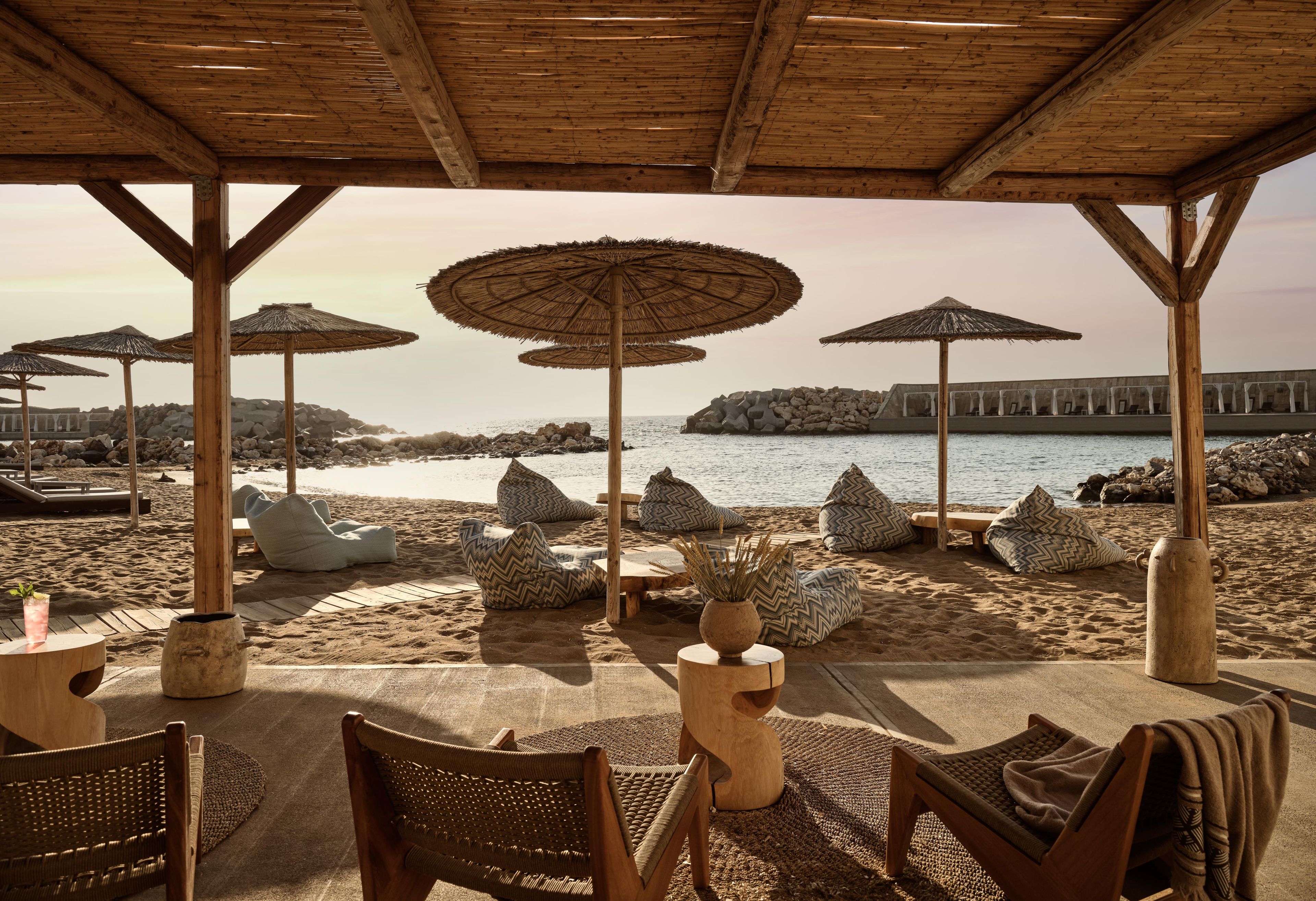 An outdoor seating area with wooden chairs and tables, surrounded by thatched umbrellas, overlooking a beach and rocky coastline.