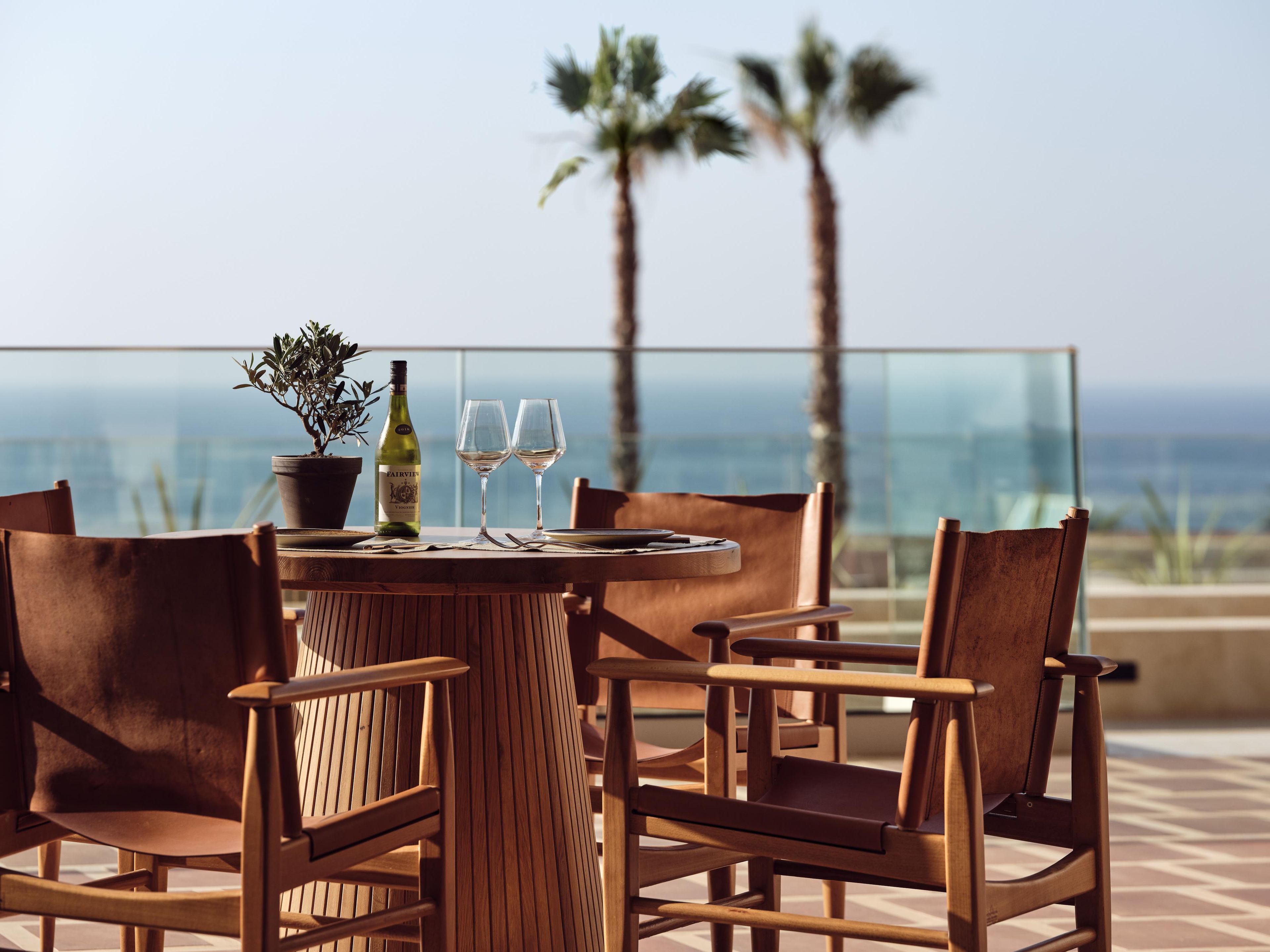 A wooden table with two wine glasses and a bottle, surrounded by chairs, overlooking a scenic view with palm trees.