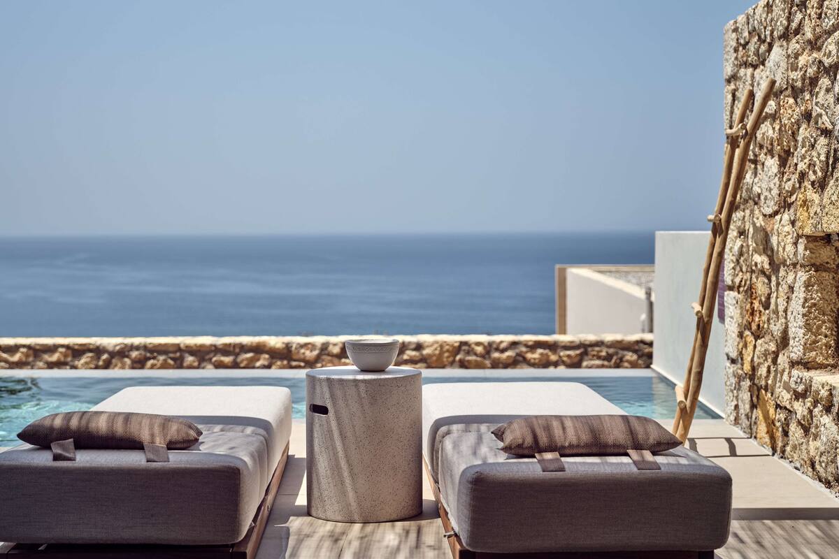 Un espacio moderno al aire libre con dos sillones acolchados y una mesita, con vista a un mar azul y despejado.