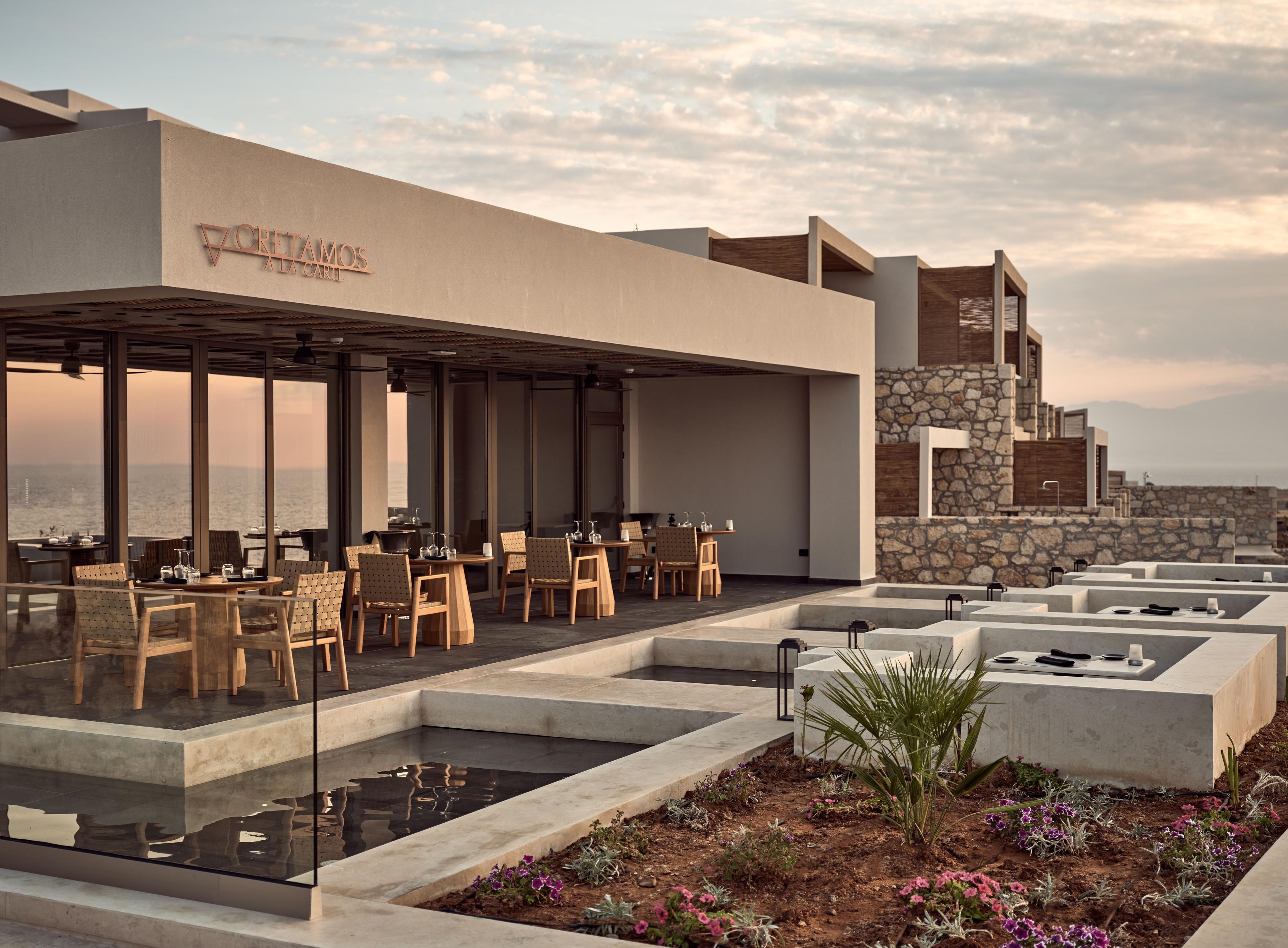 Un área de comedor exterior moderna con mesas y sillas, un muro de piedra y vista al mar.