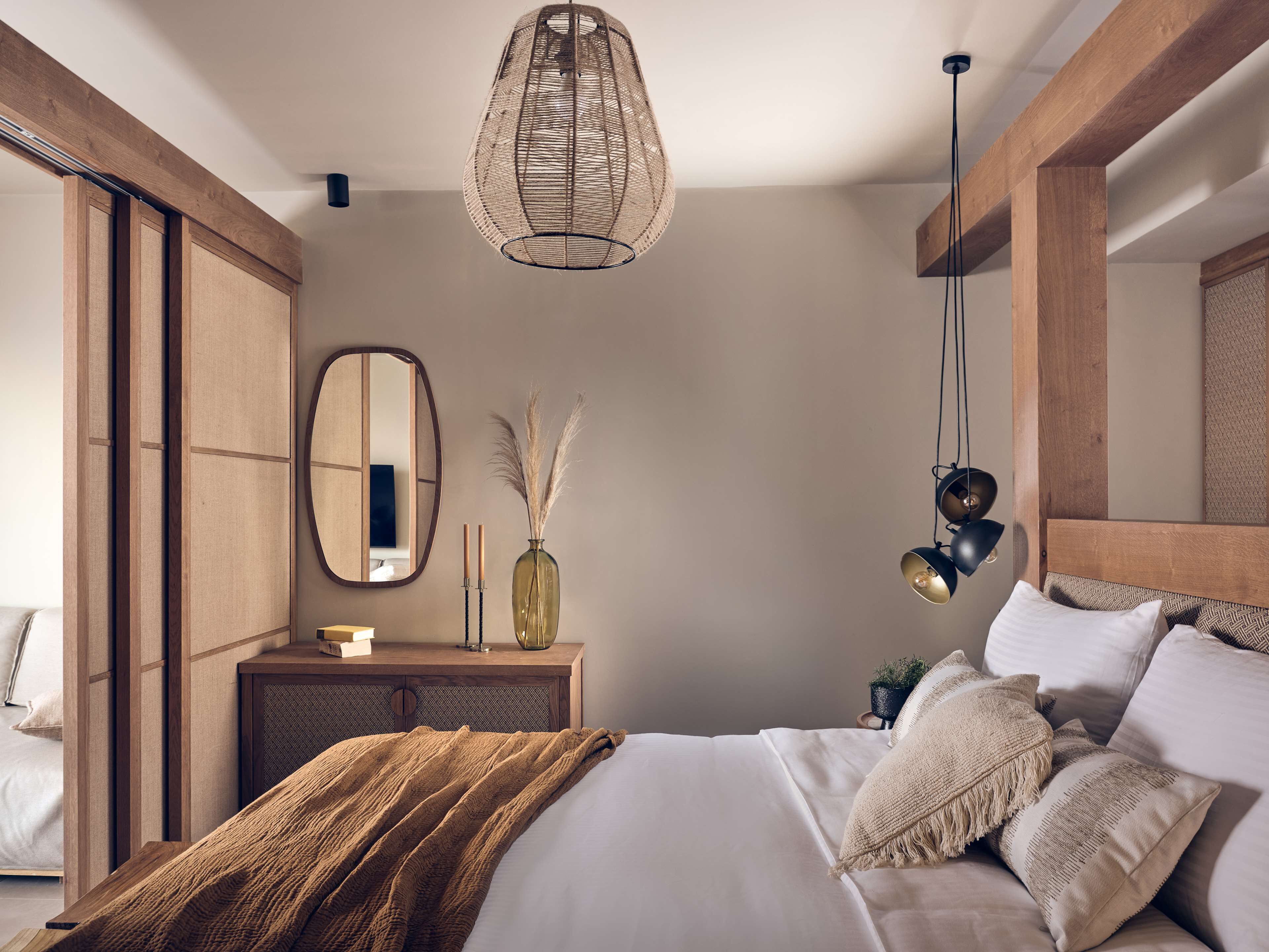 A modern bedroom with a large bed, a wooden headboard, a hanging light fixture, a mirror, and a bedside table with candles and a vase.
