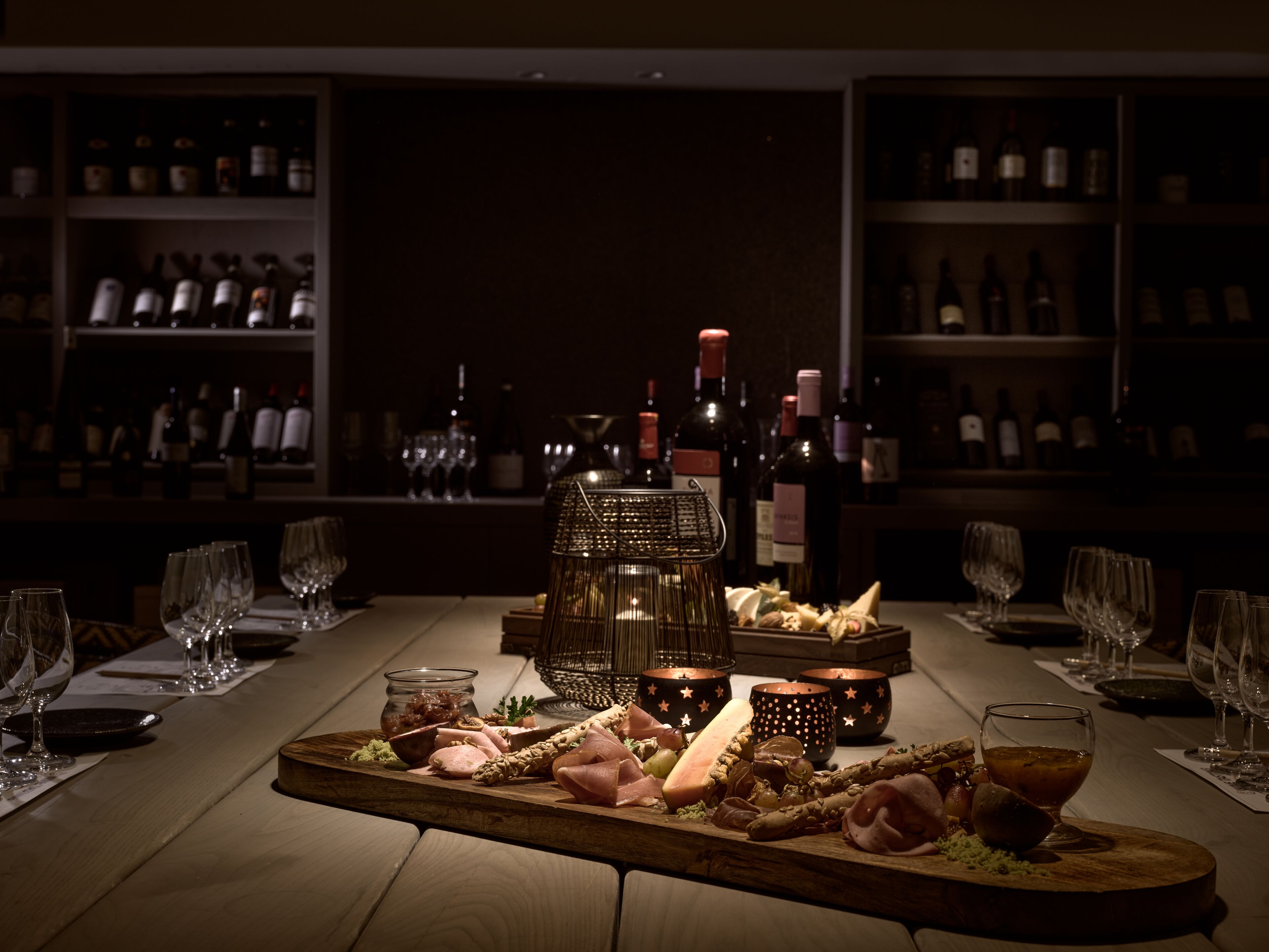 Een bar met een houten tafel waarop een vleeswarenplank, glazen en wijnflessen staan.