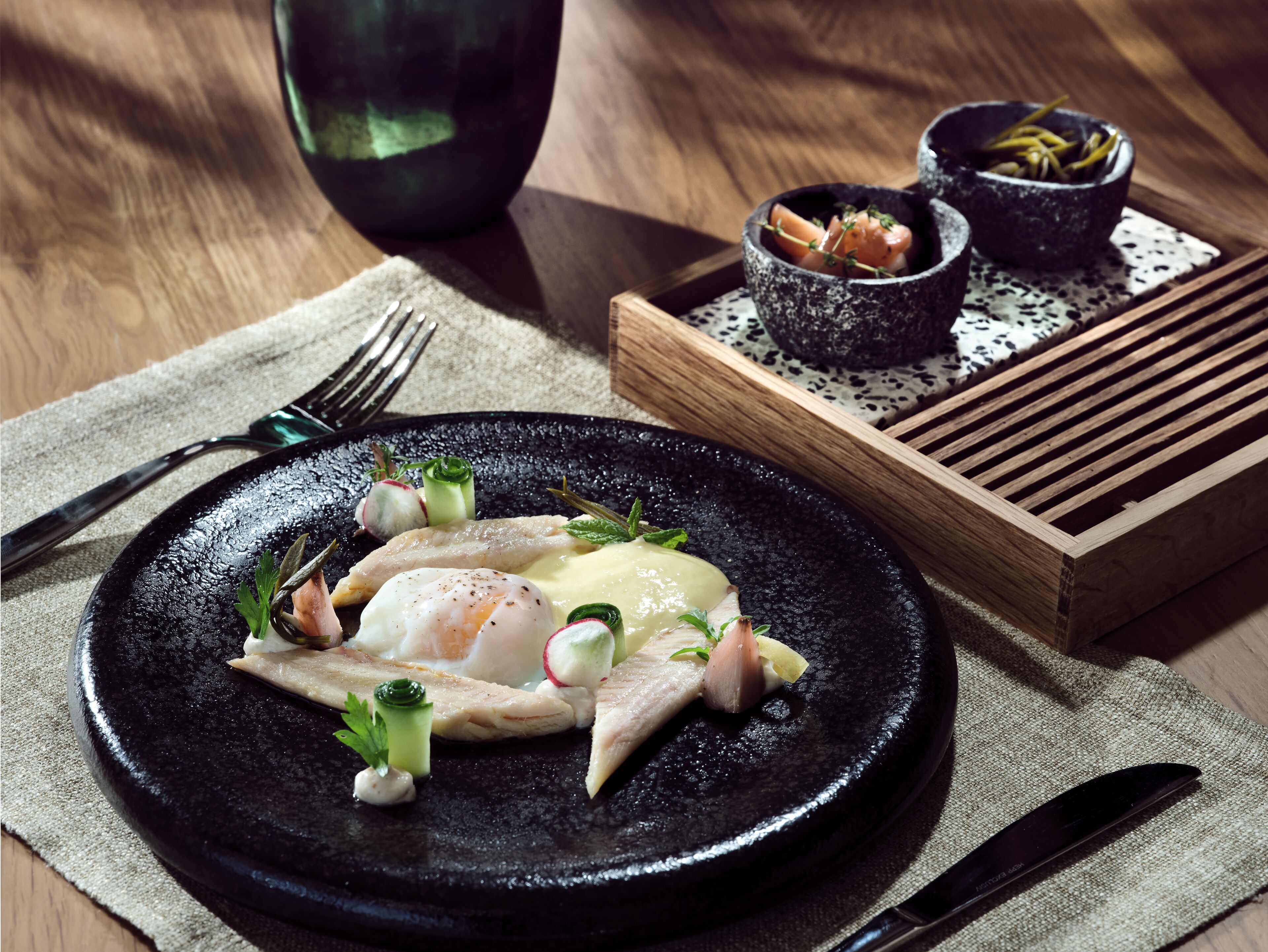 A black plate with a fish dish, garnished with herbs and vegetables, on a wooden table with a fork and knife beside it.