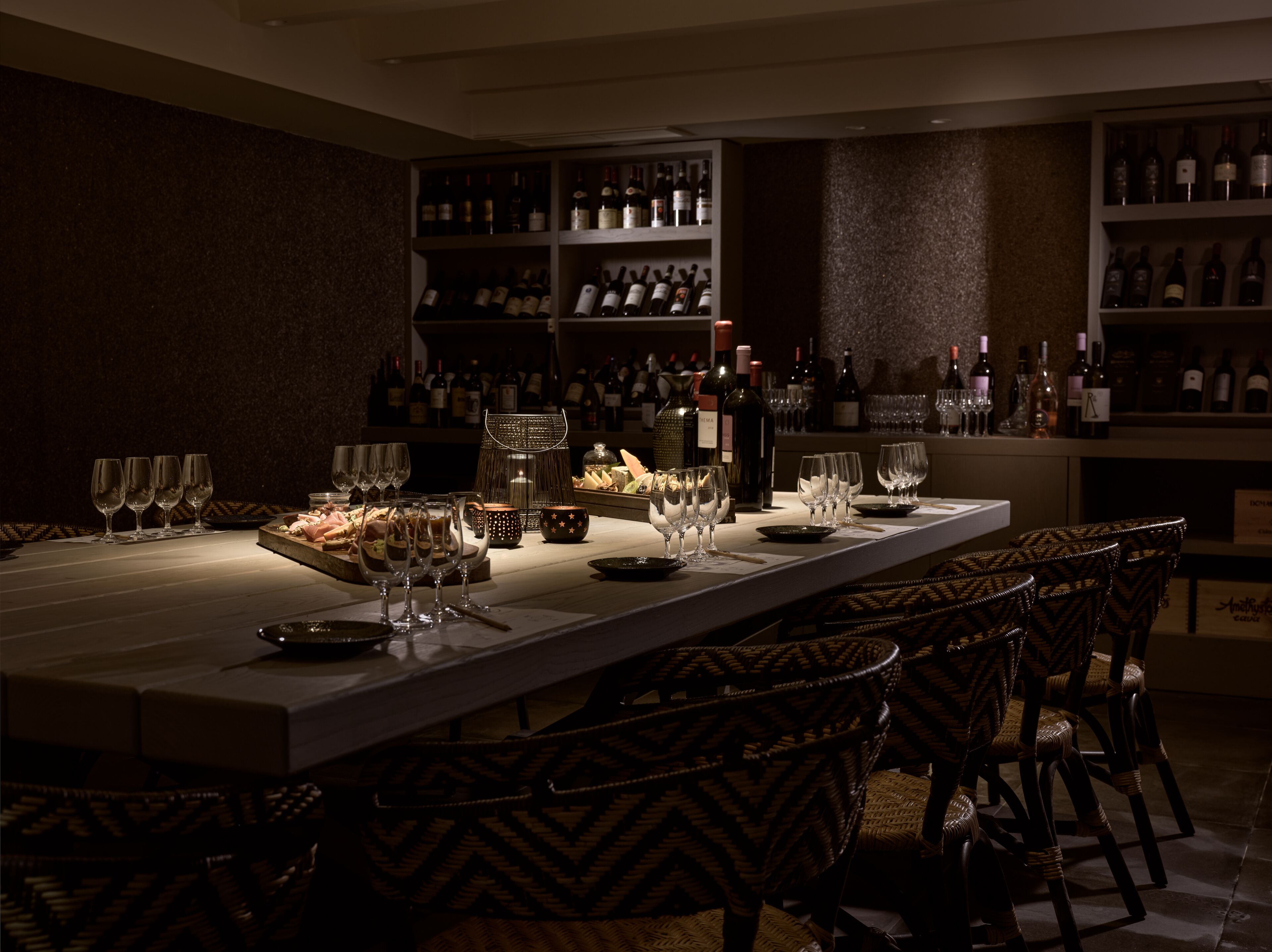 Un espace bar à la lumière tamisée, avec une longue table dressée pour le dîner, entourée de chaises en osier.