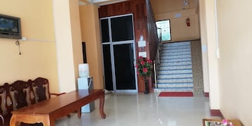 Un vestíbulo de hotel con un mostrador de recepción de madera, un amplio ventanal, una escalera y una pequeña mesa con un florero.