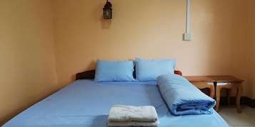 Una cama individual con ropa de cama azul, una mesita de noche pequeña y una lámpara fijada a la pared.