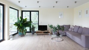 A modern living room with a large window, a flat-screen TV, a grey sofa, and several potted plants. - Immas Welt (Utting am Ammersee)