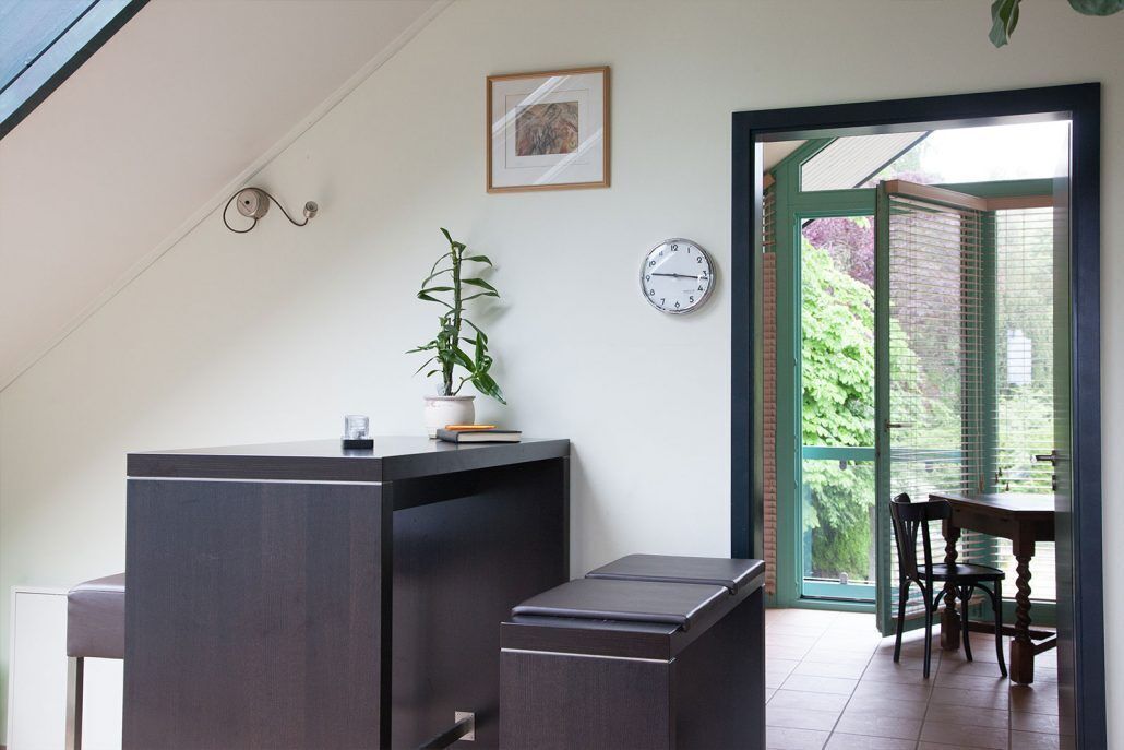 A room with a desk, a chair, a potted plant, a clock, and a door leading to an outdoor area.