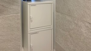 A white metal cabinet with two drawers, located in a corner with tiled walls.