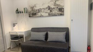 A bedroom with a bed, a desk, a chair, and a wall-mounted shelf with books and a lamp.