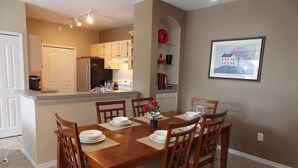 Un coin repas avec une table et des chaises en bois, une cuisine à l’arrière-plan et un tableau encadré au mur.