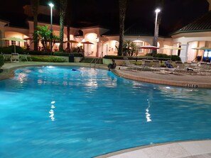 Uma piscina com espreguiçadeiras e guarda-sóis, cercada por palmeiras e um edifício com janelas em arco.