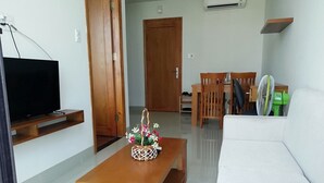 Uma sala de estar com uma TV sobre um suporte de madeira, uma mesa de centro com um arranjo floral, um sofá branco e uma área de jantar com mobiliário de madeira.
