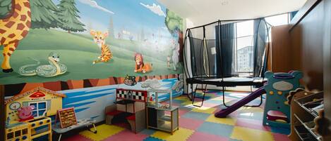 A children's playroom with colorful flooring, a mural with animals, play structures, and a window with a view.
