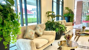 Een beige bank met bloemenkussens, een potplant en een glazen salontafel in een moderne lobby met grote ramen en een bakstenen muur.