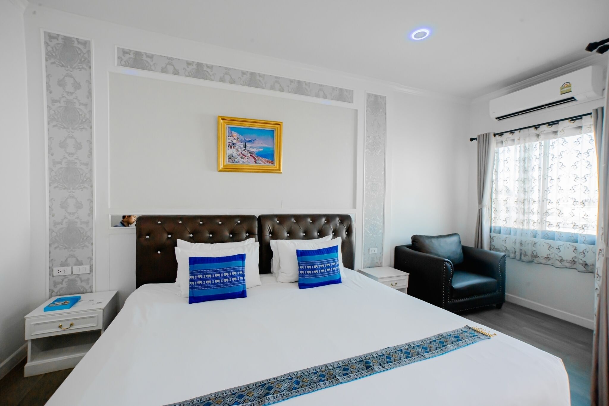 Habitación de hotel con una cama grande, cabecero, mesita de noche, una silla, ventana con cortinas y un cuadro en la pared.