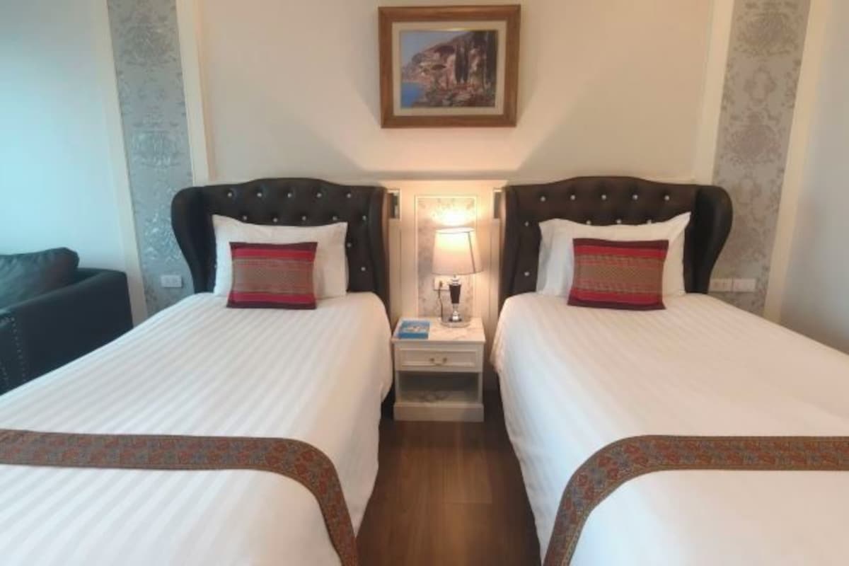 A hotel room with two beds, a nightstand with a lamp, a framed picture on the wall, and a patterned bed runner.