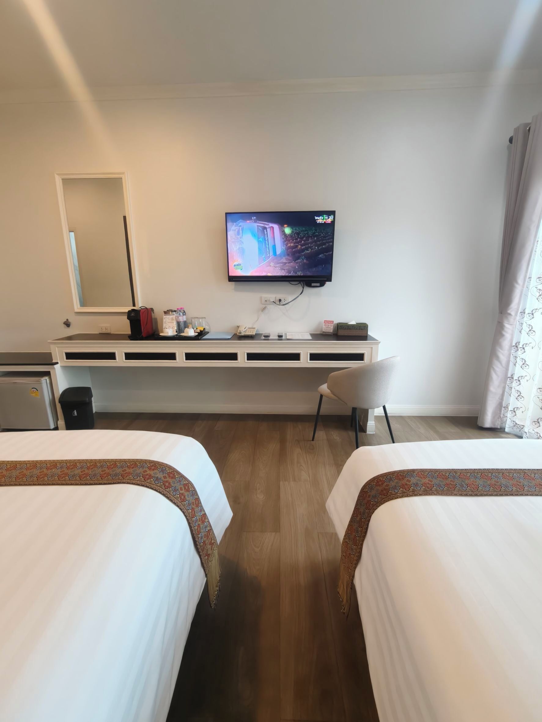 Ett hotellrum med två sängar, en tv som är monterad på väggen, ett skrivbord med en stol och en spegel.