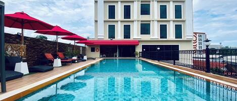 A swimming pool with lounge chairs and umbrellas, a stone wall with a mural, and a large building with multiple windows and a red awning.