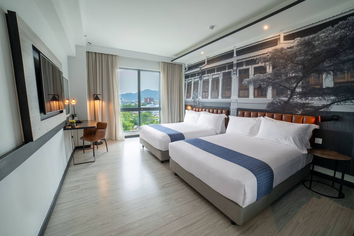 A hotel room with two beds, a desk, and a chair. There is a large window with a view of buildings and trees.