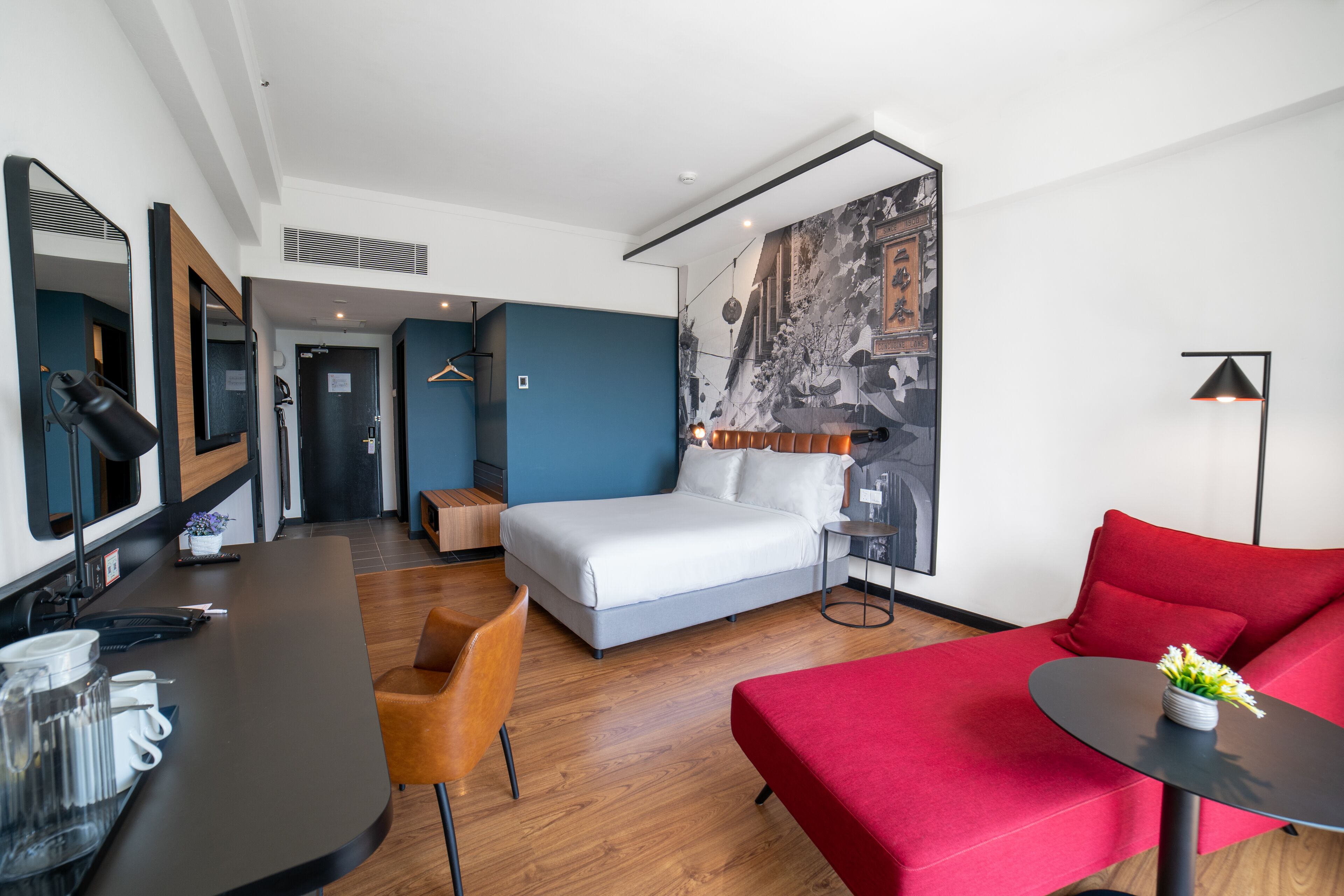 A modern hotel room with a bed, a red sofa, a small table with a vase, a desk with a lamp, and a mirror.