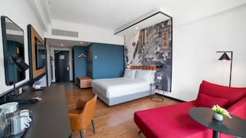 A modern hotel room with a bed, a red sofa, a small table with a vase, a desk with a lamp, and a mirror.