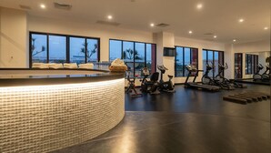 Un gimnasio moderno con un mostrador de recepción curvo, diversas máquinas de ejercicio y amplias ventanas que ofrecen vistas a palmeras y al paisaje urbano.