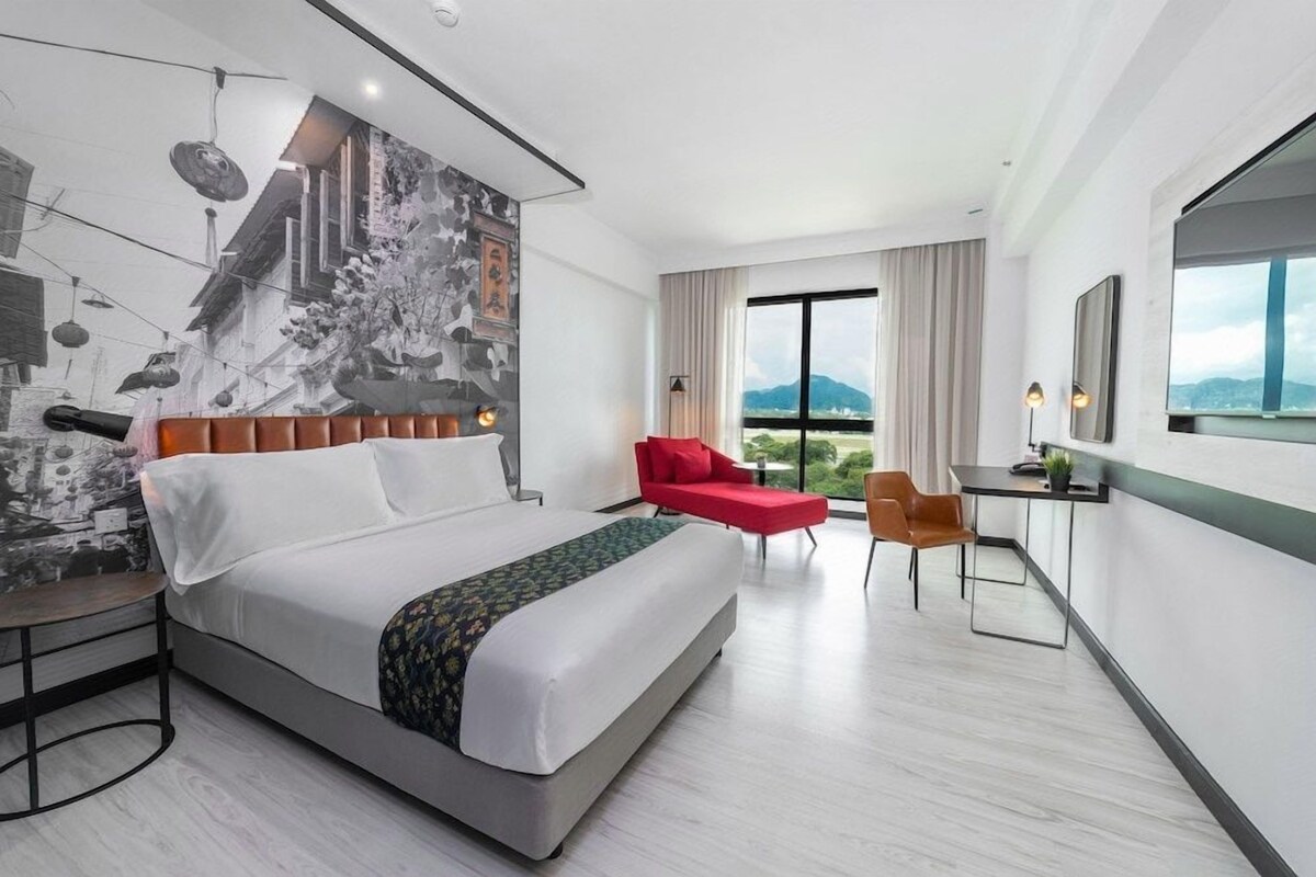 A modern hotel room with a large bed, a desk, a red chair, and a view of the outdoors.