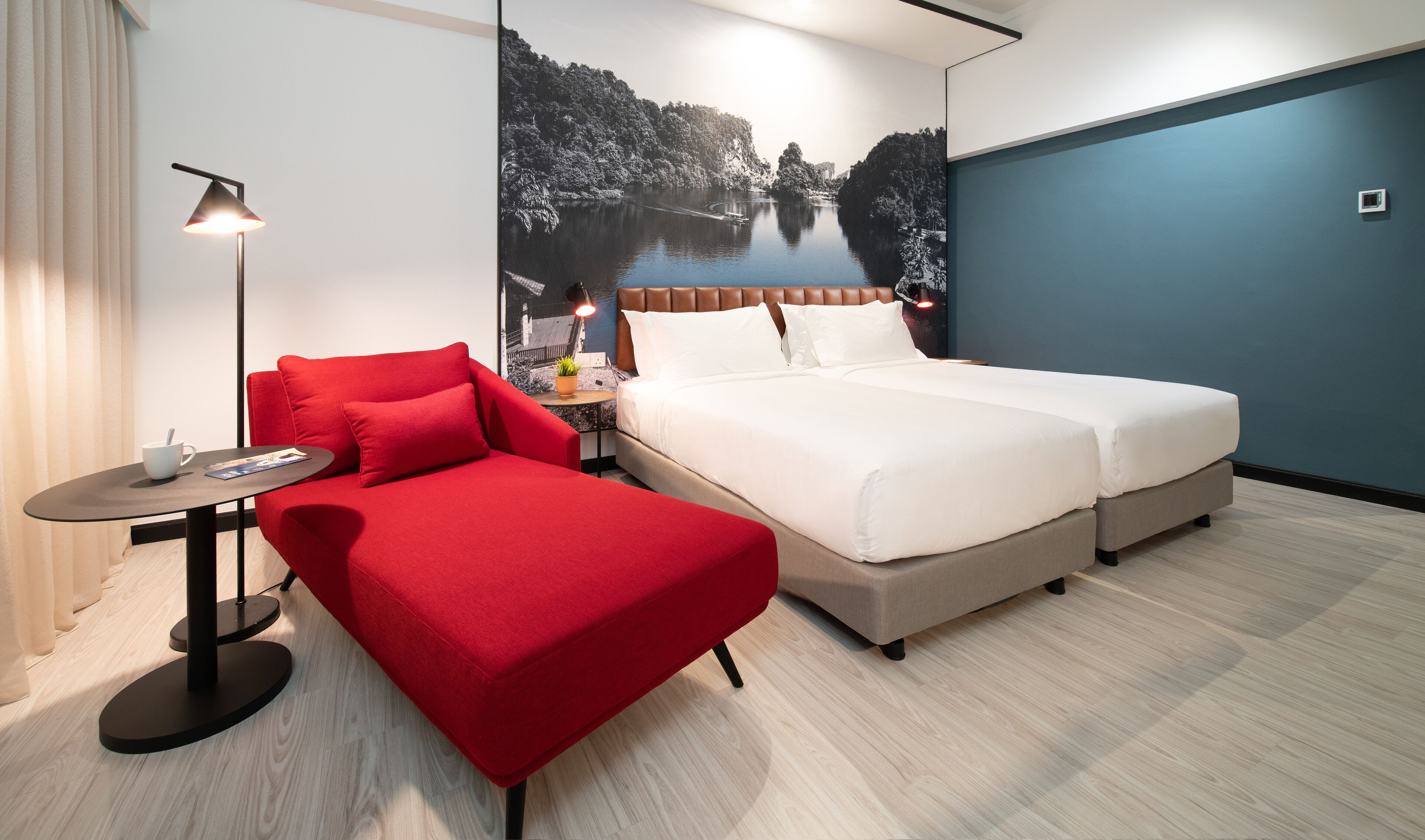 Una habitación de hotel con una cama, un sofá rojo, una mesita de noche con una lámpara y un mural grande que representa un paisaje fluvial.