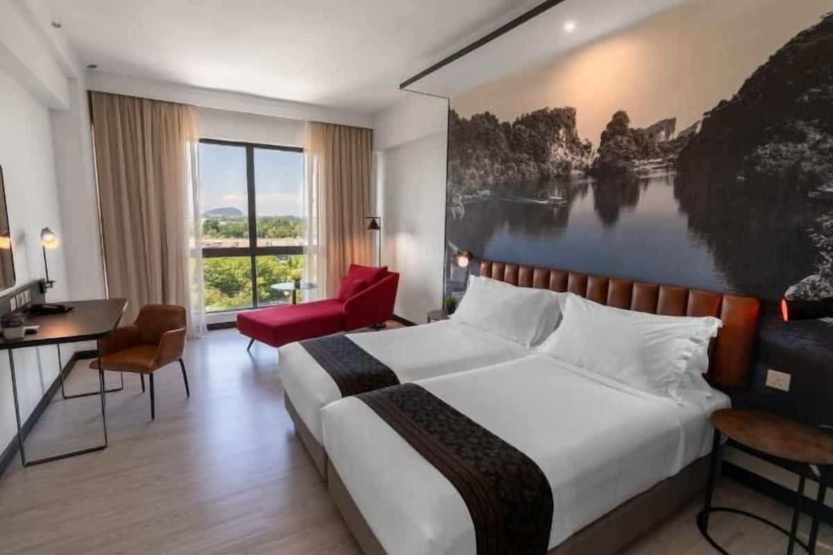 A hotel room with a bed, a desk, a chair, a red sofa, and a large window with a view of greenery.