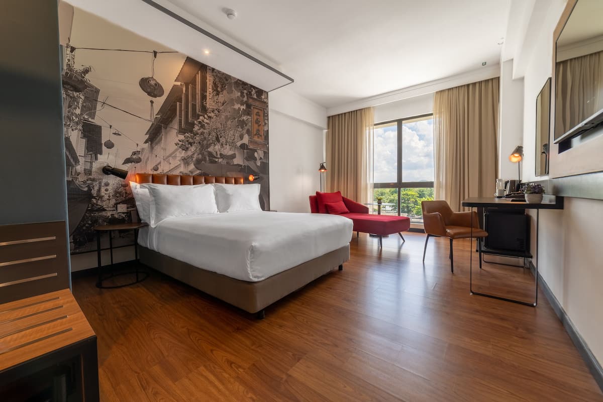 A modern hotel room with a large bed, a desk with a TV, and a view of greenery outside.