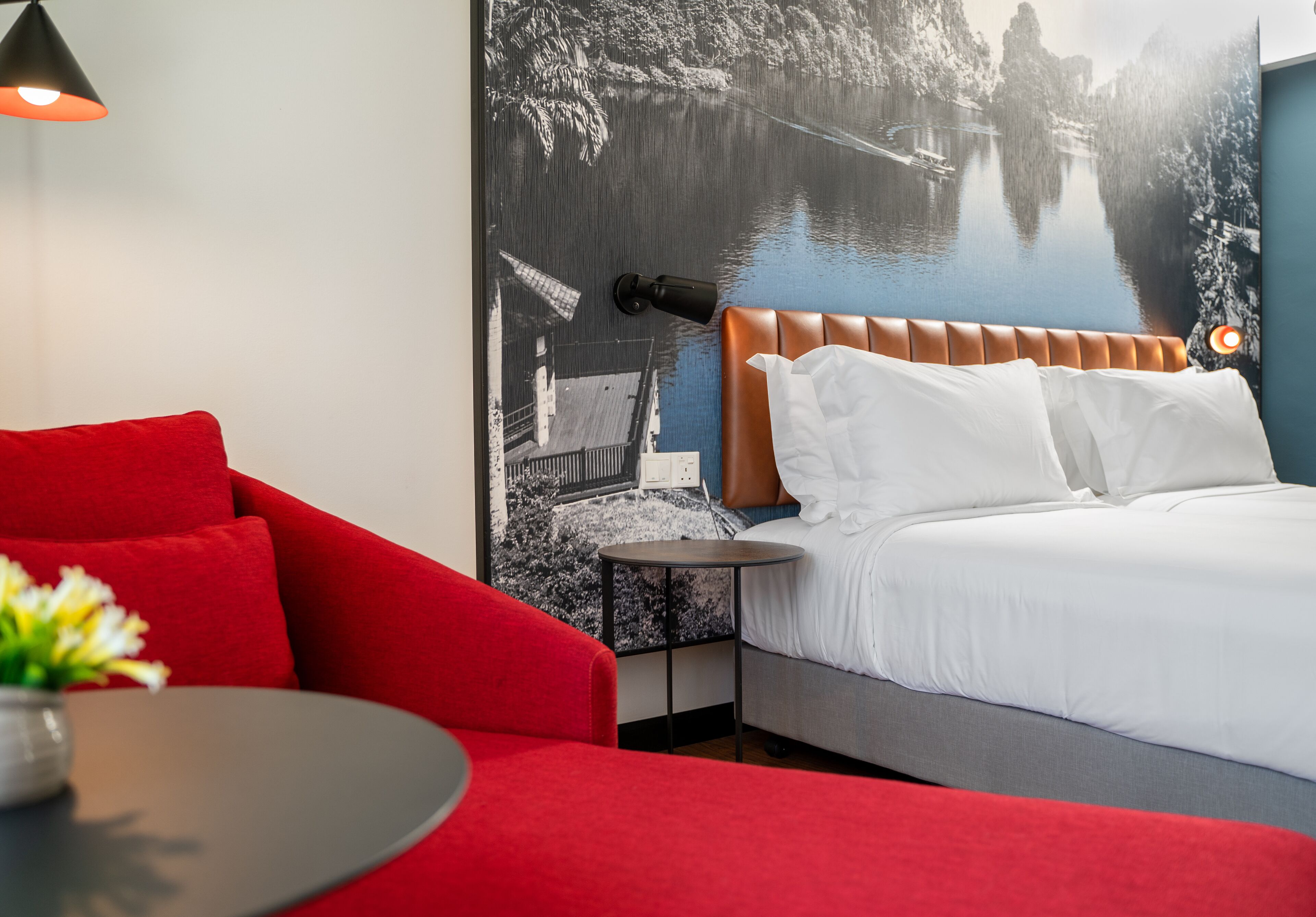 Una camera d'albergo con un letto, una poltrona rossa, un tavolino e una lampada a parete.