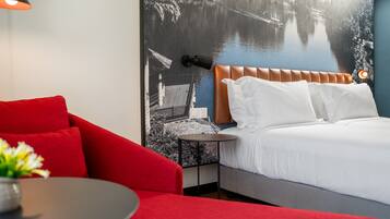 A hotel room with a bed, a red armchair, a small table, and a wall-mounted lamp.