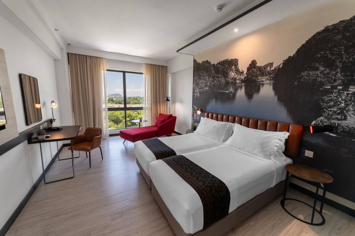 A modern hotel room with a large bed, a desk, a red chair, and a view of a lake through the window.