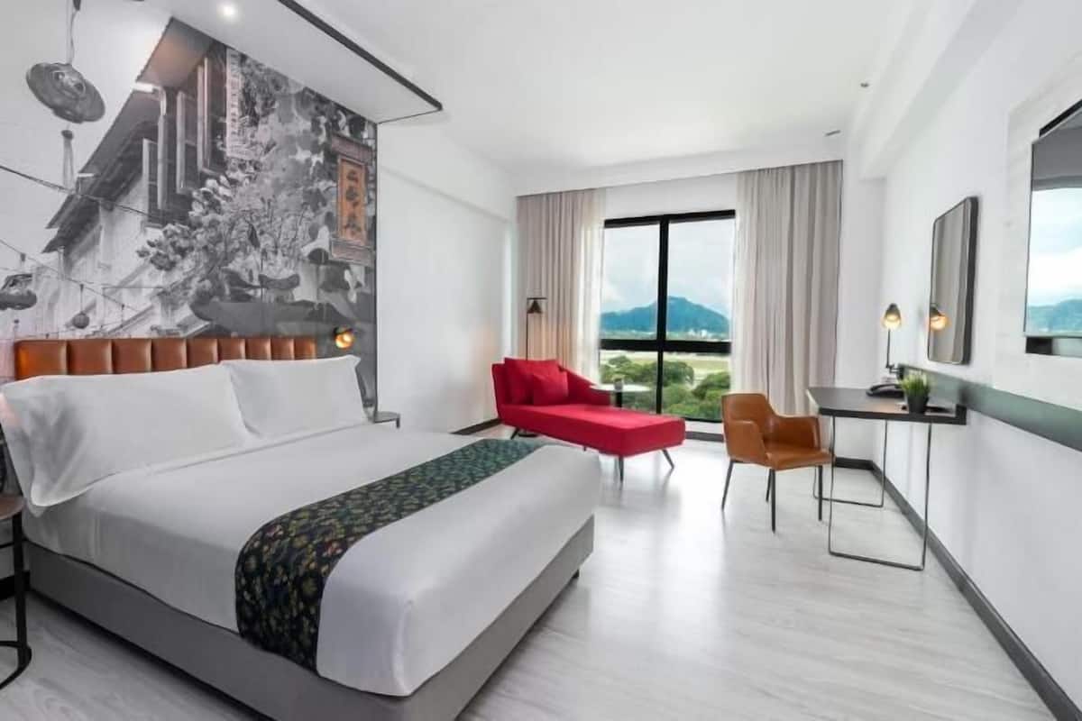 A modern hotel room with a large bed, a desk, a red chair, and a view of the outdoors.