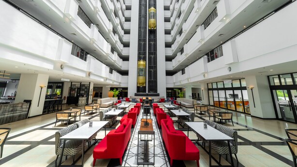 Un espacio interior amplio con varias mesas de comedor y asientos rojos, un elemento vertical central, y un techo alto con luminarias.