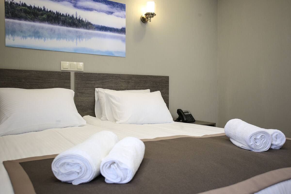 A hotel room with a large bed, two rolled towels, a phone on the nightstand, and a painting of a forest on the wall.