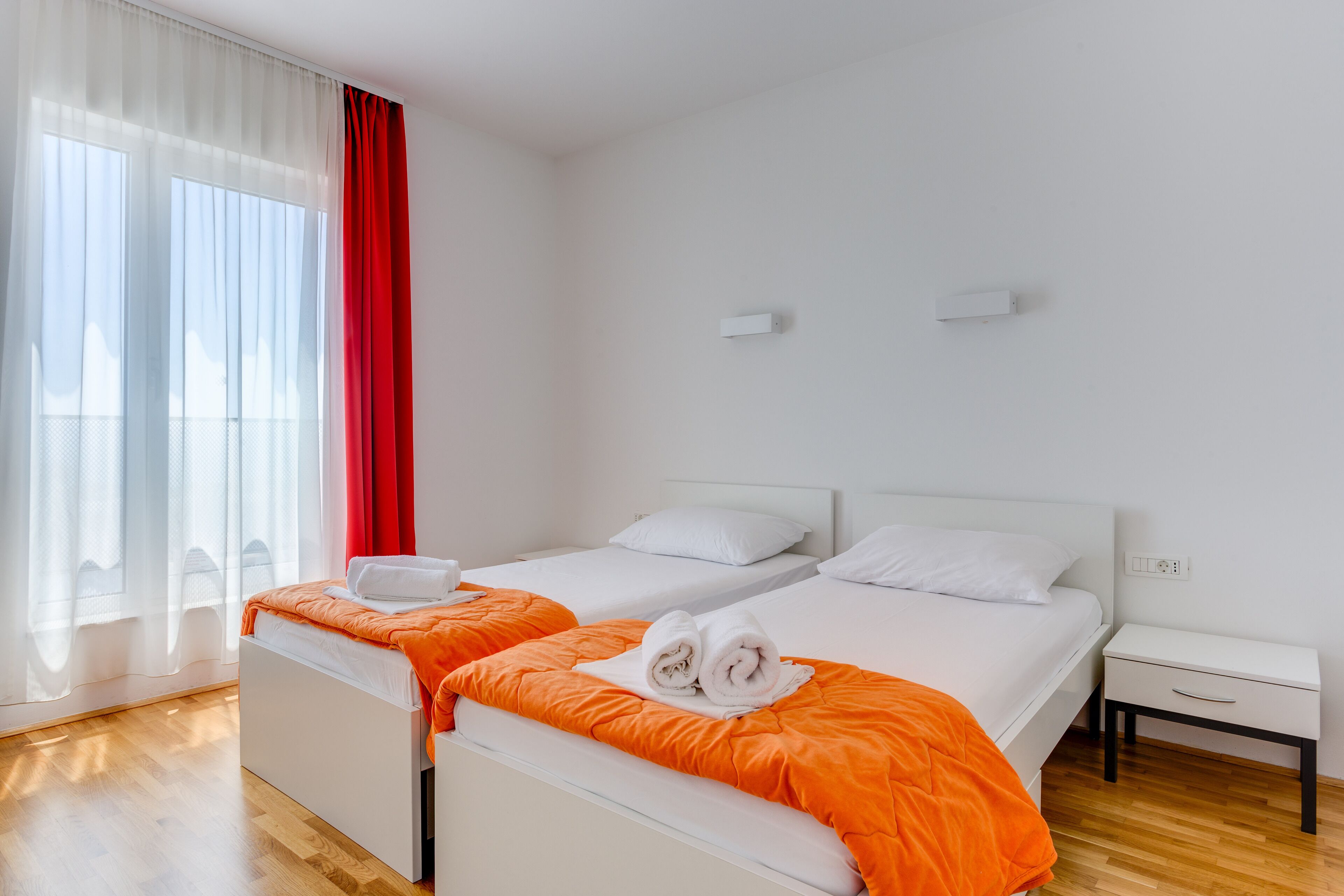 A hotel room with two beds, white bedding, a wooden floor, a red curtain, and a white nightstand.