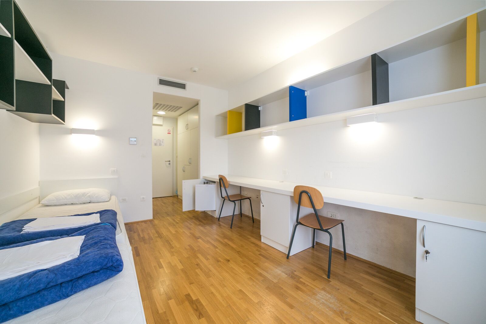 A dormitory room with two beds, a desk, chairs, and built-in shelving.