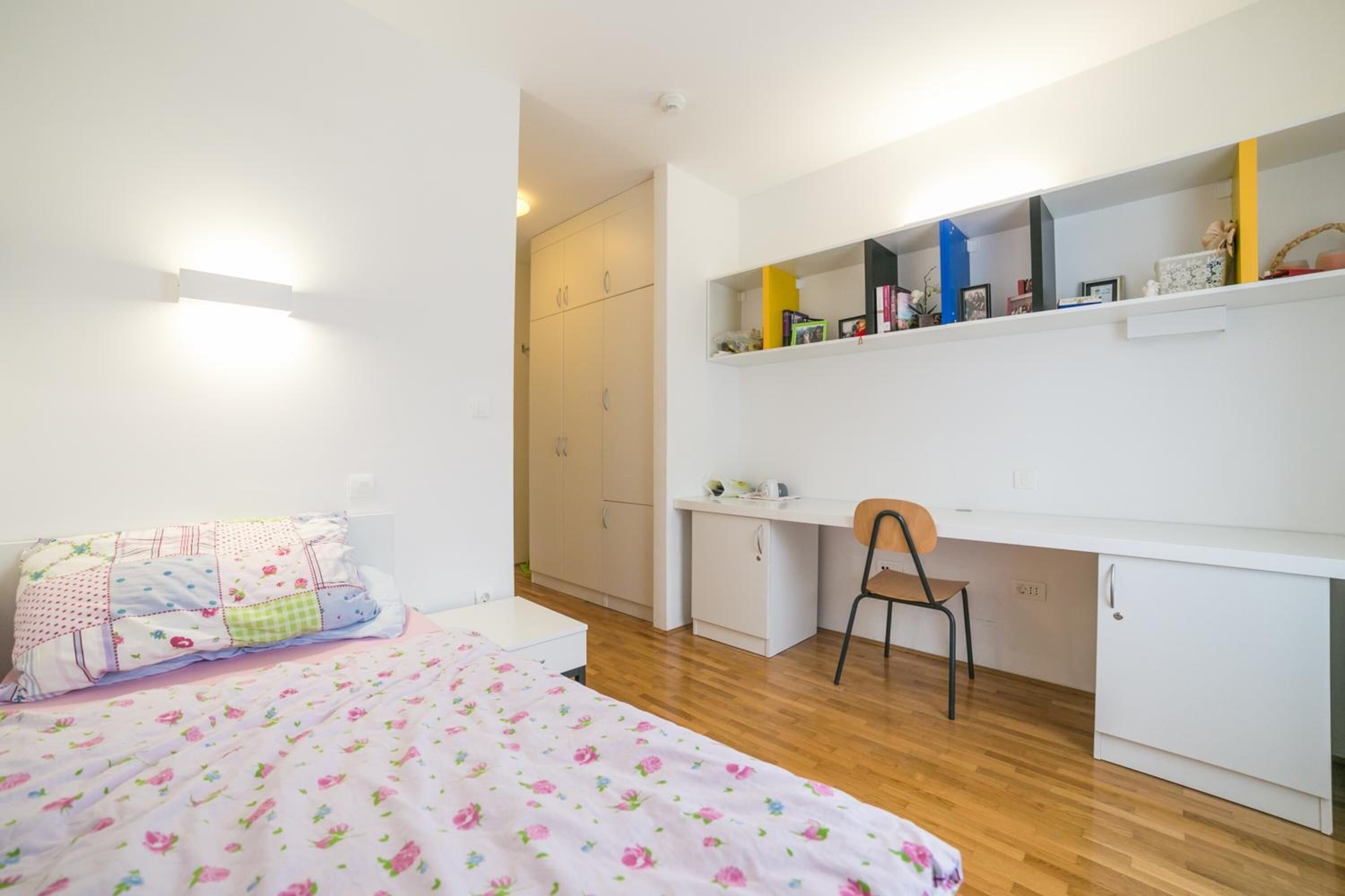 Un dormitorio con una cama, un escritorio y una silla. Hay un armario y una estantería con diversos objetos.